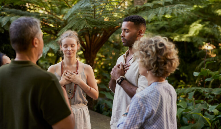 Group of multiethnic adults standing peacefully in circle with hands on their hearts during a wellness retreat in nature. Diverse people practice mindfulness and gratitude together in a forest. Mixed race group sharing a meditation on fourth chakra, heart chakra, outdoor with copy space.