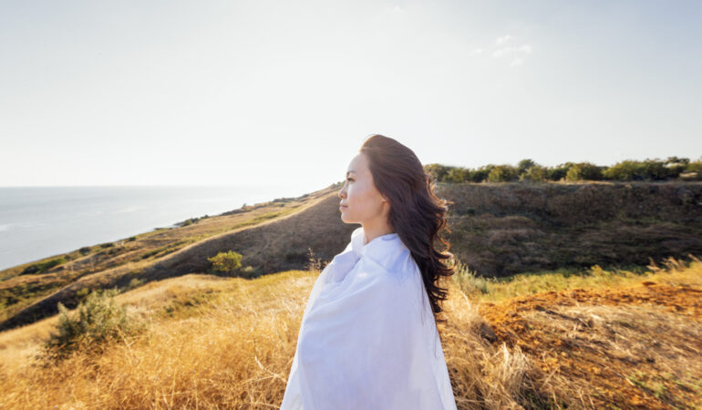 A young handsome Asian woman wrapped in a white sheet and looks at the seascape. A charming Korean woman enjoys nature. Profile of an attractive girl in the rays of the sun at sunset outdoors.