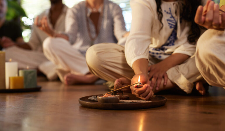 Close up hand of young woman lighting insence stick while practicing meditation. Detail of a woman lighting incense stick with lighter before meditation ritual at indoor wellness retreat. Spiritual ceremony begins with sacred fire and intention.