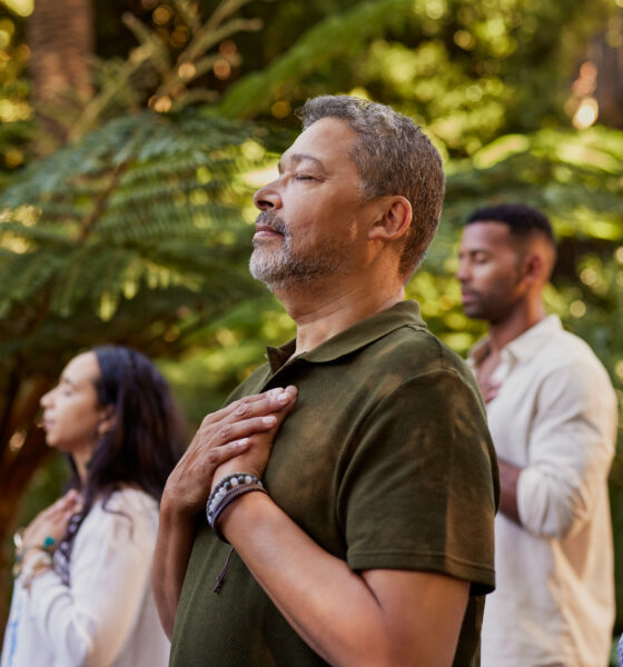Group of multiethnic people standing outdoors with eyes closed and hands on their hearts during a meditation session. Multiracial adults practicing gratitude and mindfulness together. Mature latin man connecting to inner peace and presence at wellness retreat center in nature.